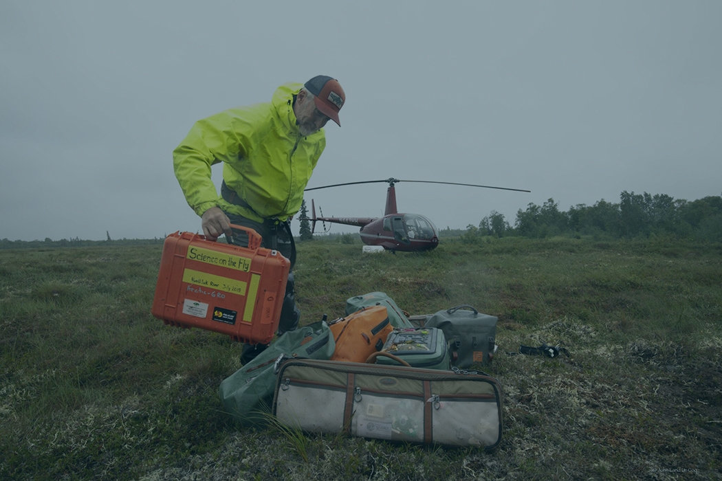 Max Holmes collects equipment used for Arctic research with helicopter in background.